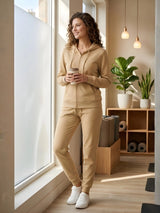 Woman in beige tracksuit standing by a window holding a coffee cup in a modern room.