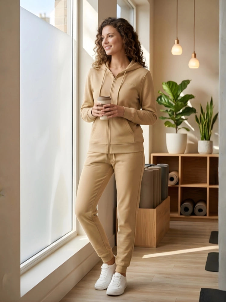 Woman in beige tracksuit standing by a window holding a coffee cup in a modern room.