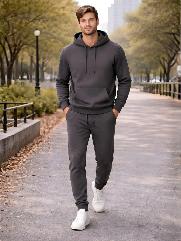 Man wearing a gray hoodie and jogger pants walking on a path with trees and buildings in the background. 

Dark grey men’s pullover hoodie with matching jogger trousers by Stylewar