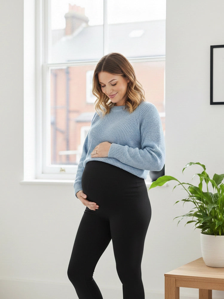Pregnant woman wearing a light blue sweater and black leggings indoors.