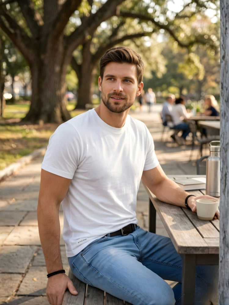 Man sitting at a wooden table outdoors with trees and people in the background.

Men’s white crew neck t-shirt by Stylewar