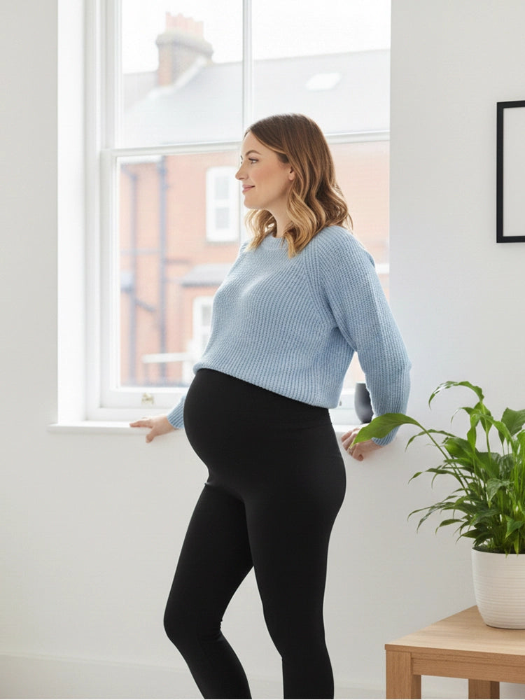 Woman wearing a light blue sweater and black leggings standing indoors near a window.